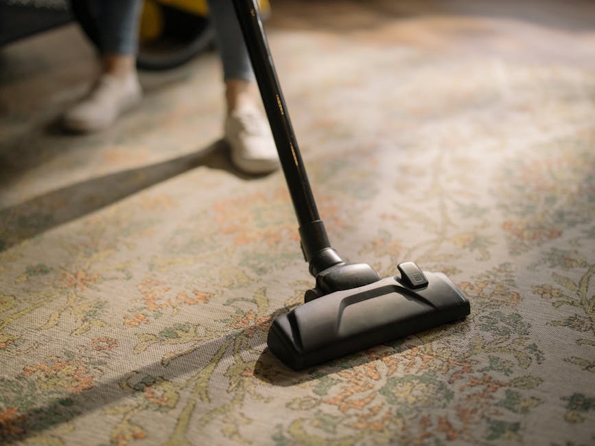 A person kneeling on a patterned carpet in a domestic setting, using a handheld vacuum cleaner with a yellow and black body to perform surface cleaning and deep cleaning of the carpet. The vacuum's du
