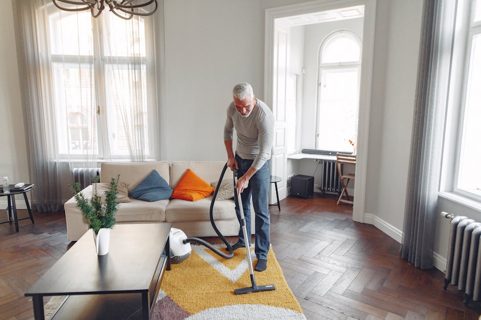 A woman with brown hair tied back, wearing a light-colored striped shirt and jeans, is kneeling on an ornate patterned rug in a living room. She is using a blue cloth to clean or polish the dark woode